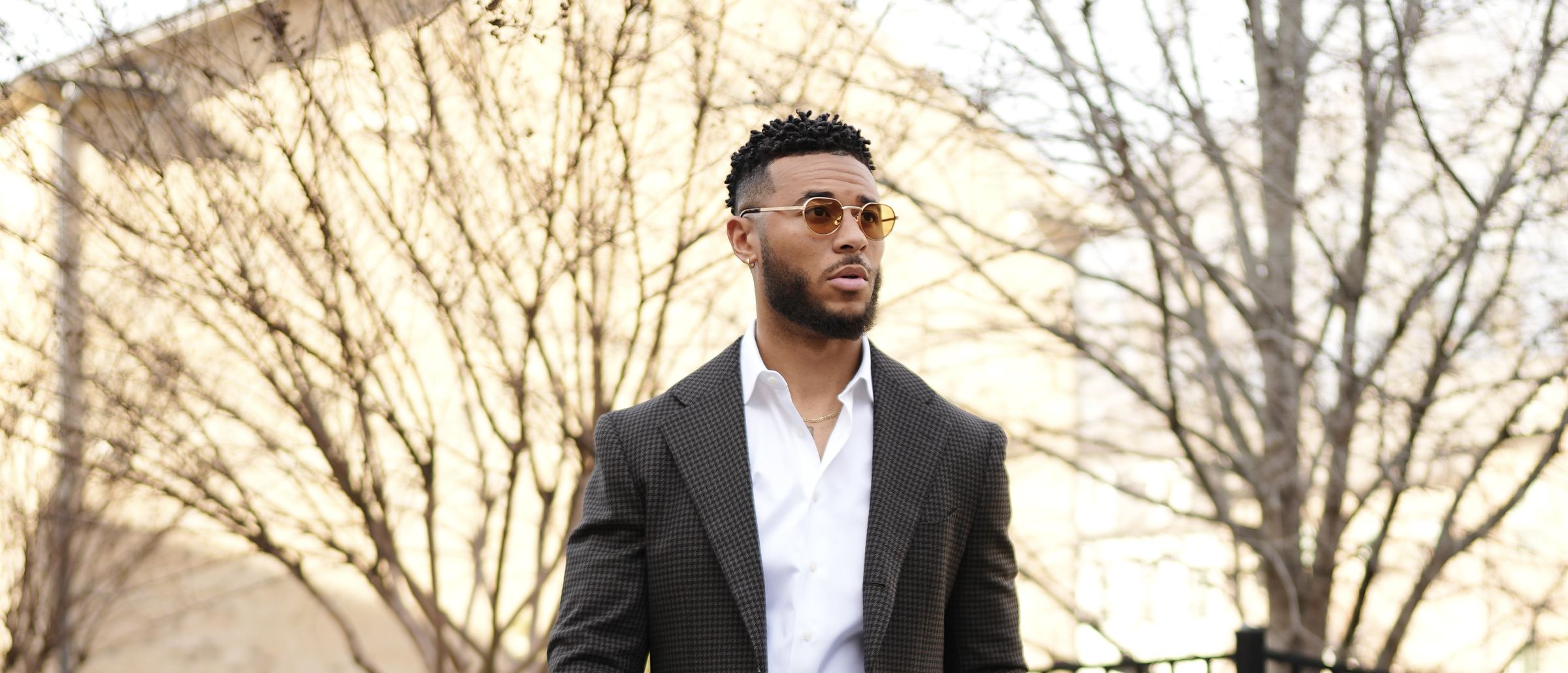 Trey Bryant in a houndstooth jacket and a white shirt, wearing tinted sunglasses, under bare trees